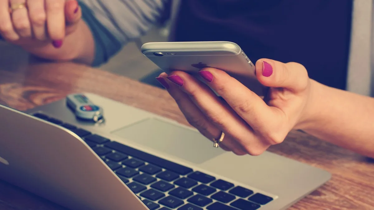 A close-up of hands with pink nail polish holding a smartphone over an open laptop on a wooden table.