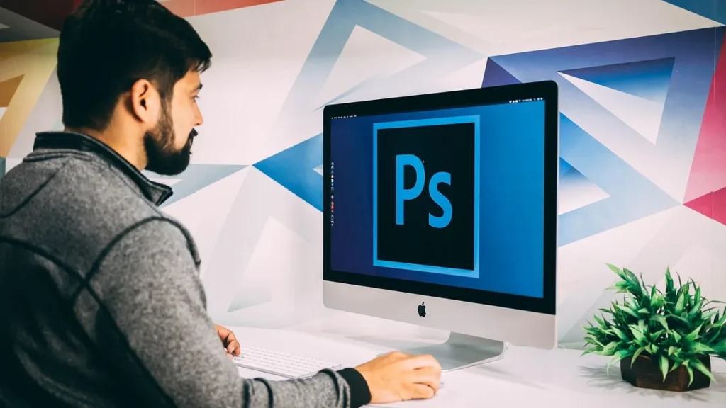 A man in profile working on an iMac displaying the Adobe Photoshop logo against a colorful geometric wall.