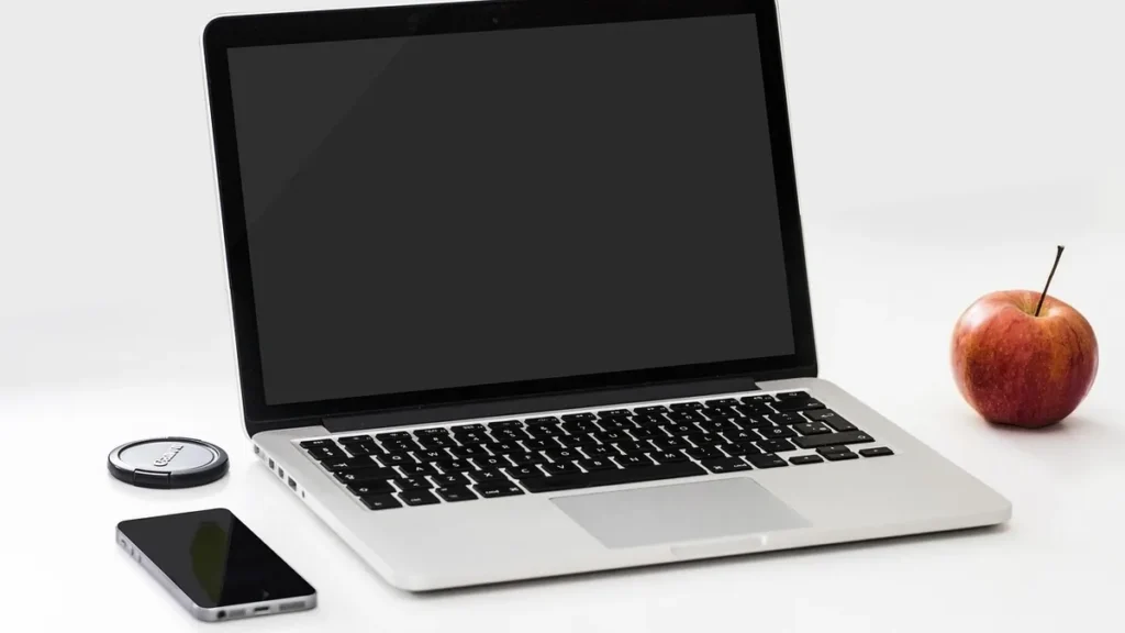 A silver laptop with a black screen, a smartphone, a Canon lens cap, and a red apple on a white surface.