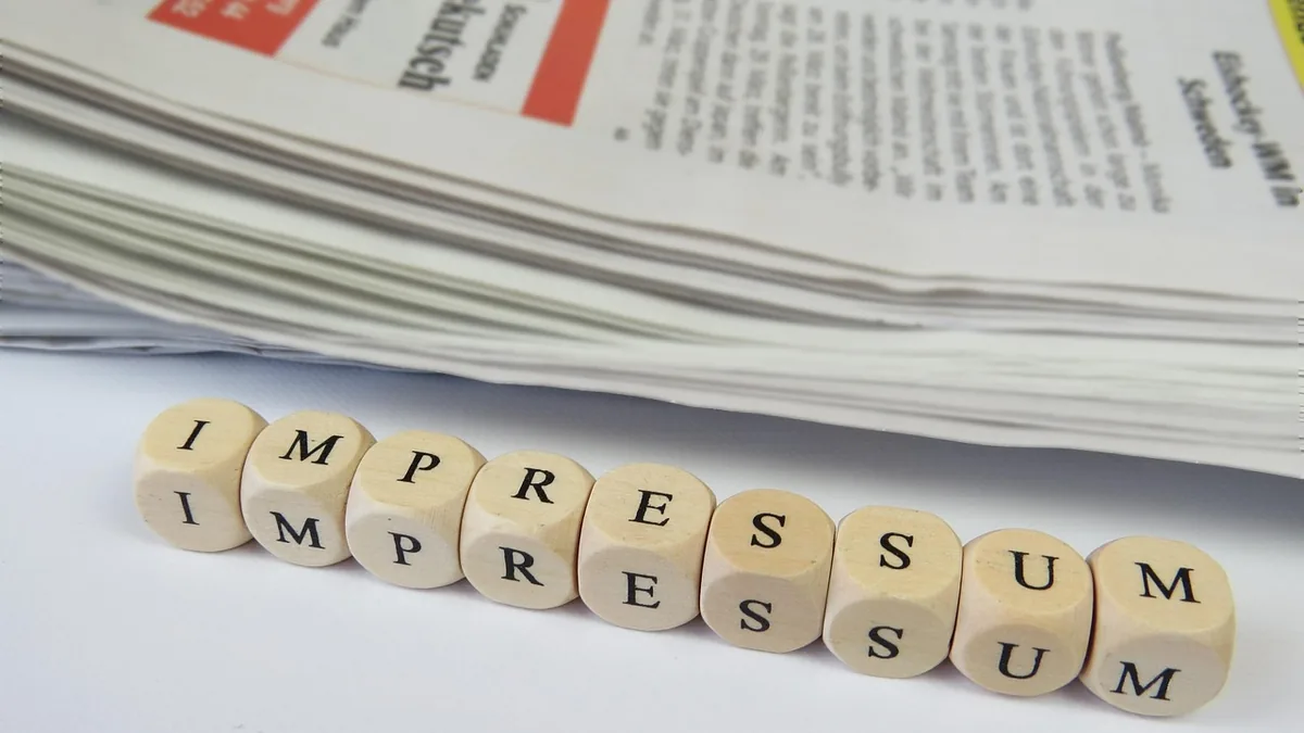 Wooden blocks spelling IMPRESSUM in front of a stack of newspapers, symbolizing legal disclosure.