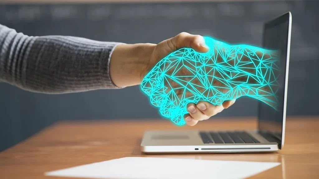 A glowing blue digital hand from a laptop shakes a human hand, symbolizing AI integration and cybersecurity.