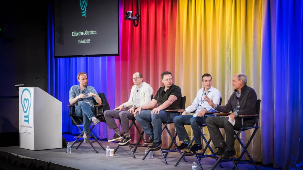 Panelists, including Elon Musk, on stage at the Effective Altruism Global 2015 event with colorful curtains.