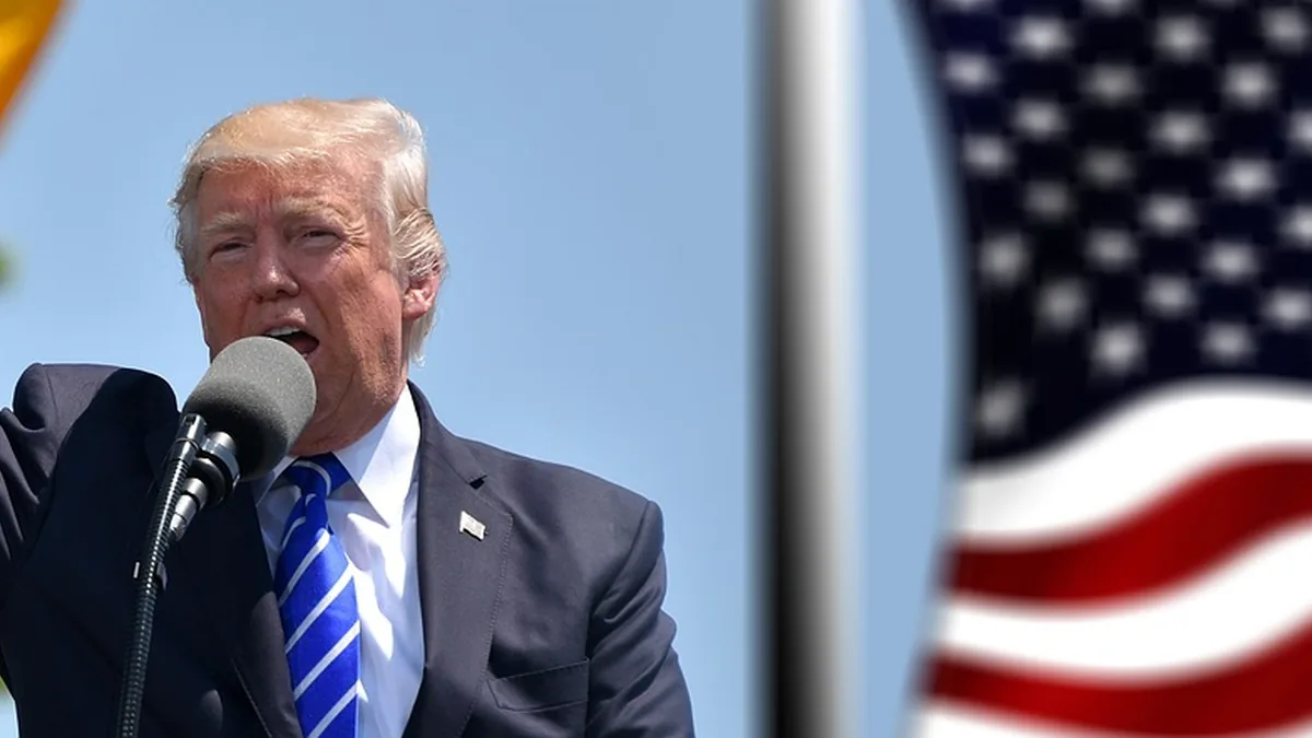 Donald Trump speaking into a microphone with a blurred American flag in the background under a blue sky.
