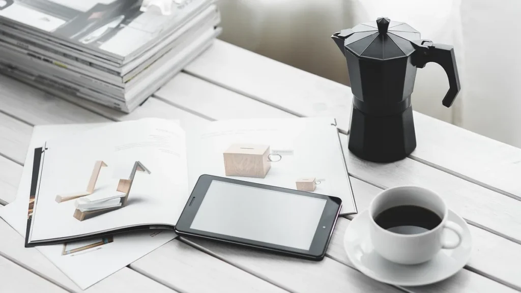 A black tablet, a cup of coffee, and a black moka pot on a white wooden table with magazines.