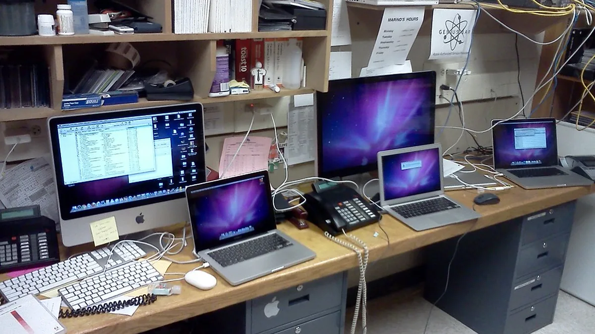 Multiple Apple desktop and laptop computers are cluttered on a wooden desk in an IT workspace.