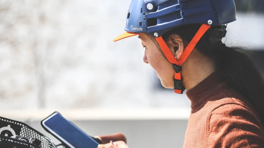 Profile of a woman wearing a blue Overade helmet with orange straps looking down at a smartphone.