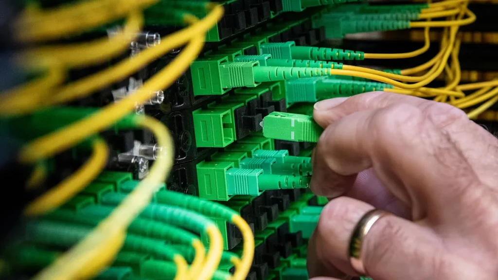A close-up of a technician's hand plugging green fiber optic cables into a network patch panel with yellow cables.