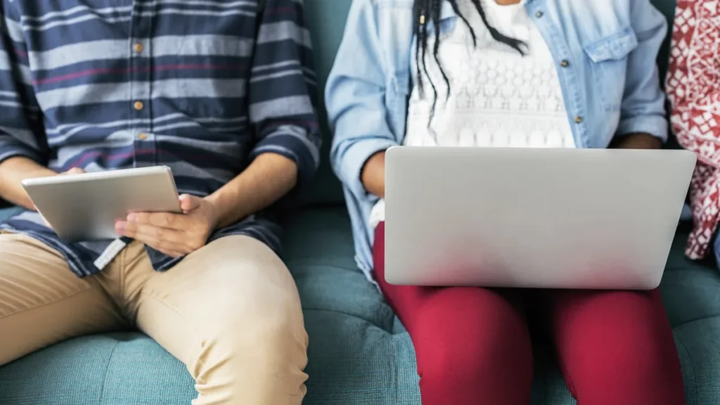 People sitting on a couch using a tablet and a silver laptop, cropped torsos.