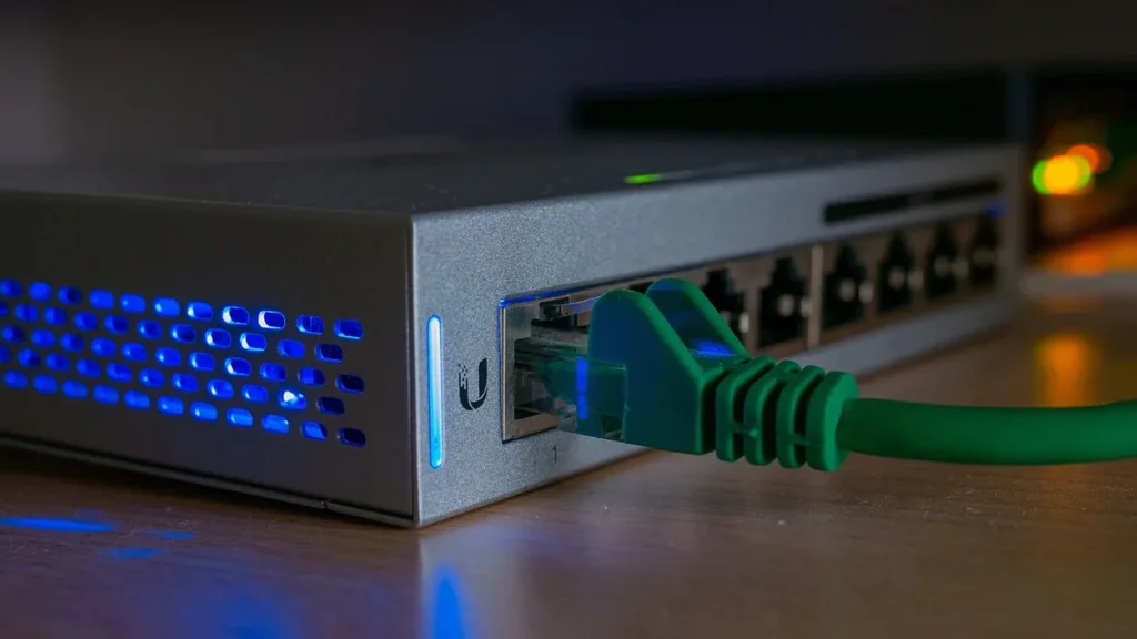 Green ethernet cable plugged into a silver network switch with glowing blue LEDs.