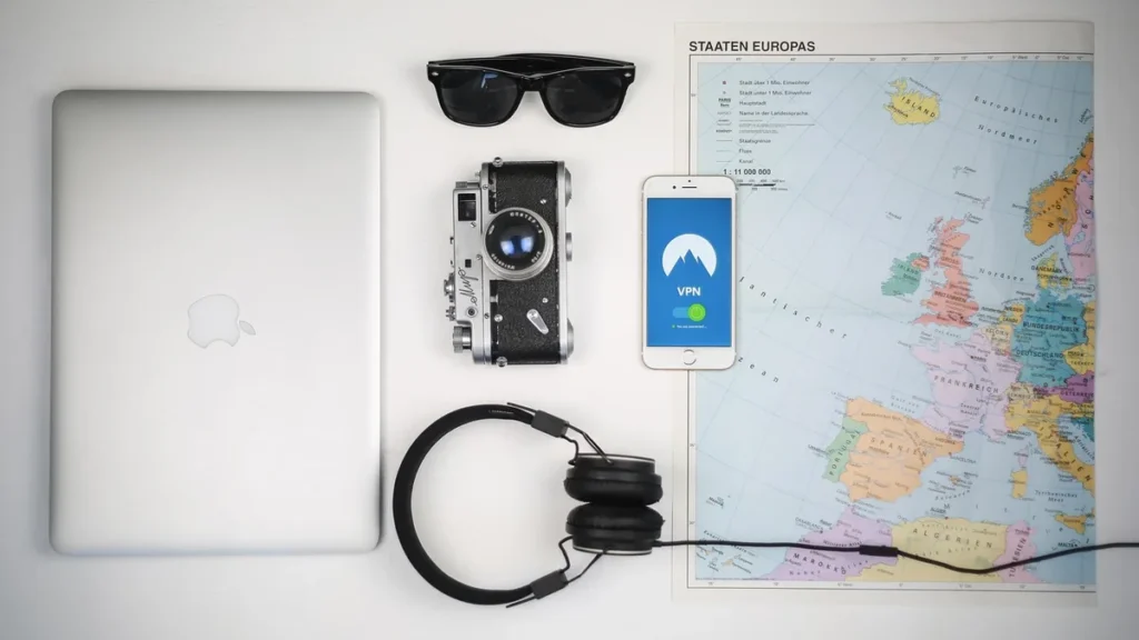 Flat lay of a laptop, camera, smartphone with VPN app, sunglasses, headphones, and a map of Europe.