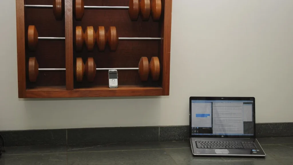 A laptop on the floor below a giant wooden abacus with a small mobile phone on the shelf.