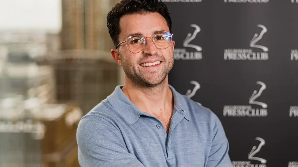 Smiling man in glasses and a blue polo shirt stands before a Melbourne Press Club backdrop.