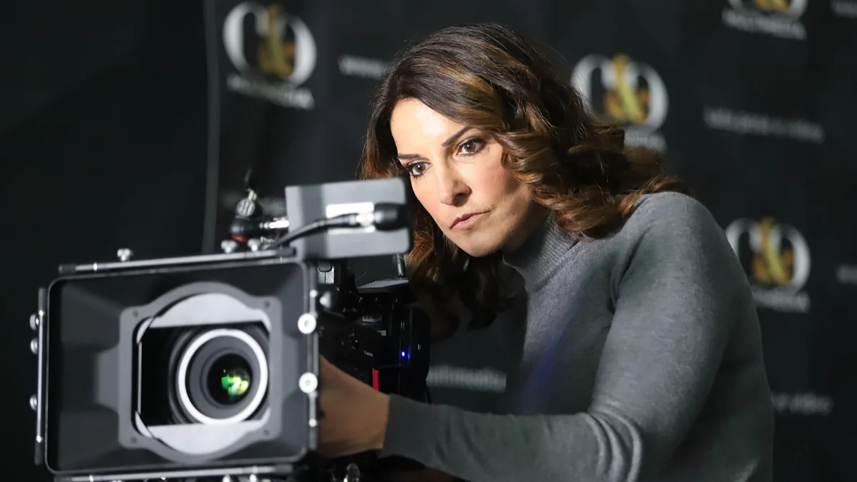 A woman in a grey turtleneck looks intently at a large professional video camera against a dark branded backdrop.