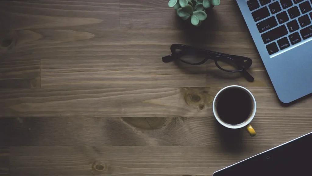 Top-down view of a wooden workspace with a laptop, coffee cup, glasses, and a small succulent plant.
