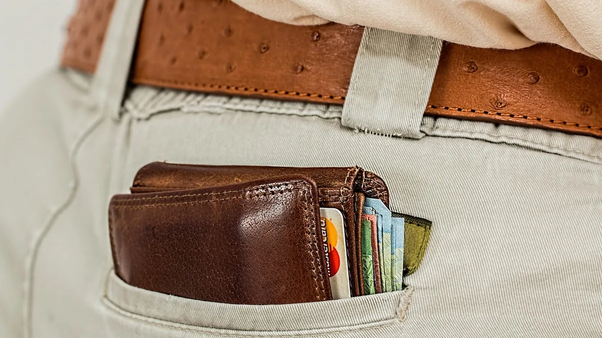 A brown leather wallet filled with cash and cards protruding from the back pocket of light-colored trousers.