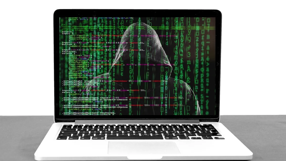 Laptop screen displaying a hooded hacker silhouette overlaid with green matrix-style code and CSS script.