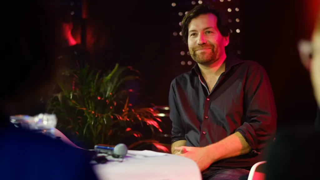 A man in a dark shirt smiles while sitting on a panel under red and dark stage lighting, with a plant in the background.