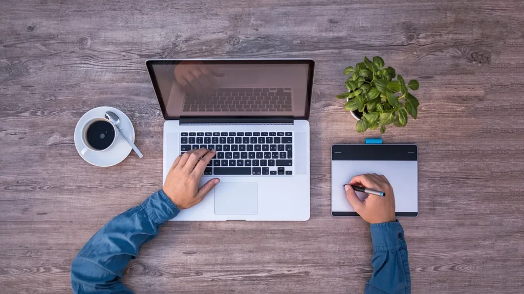 Top-down view of hands on a laptop and drawing tablet on a wooden desk with coffee and a plant.