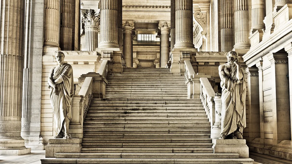 Grand classical staircase flanked by two marble statues of ancient philosophers amidst towering columns.