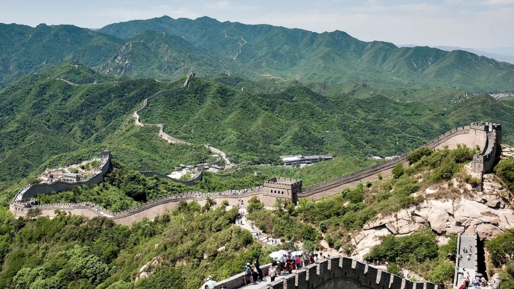 The Great Wall of China winds through lush green hills, with tourists walking along the ancient stone path.
