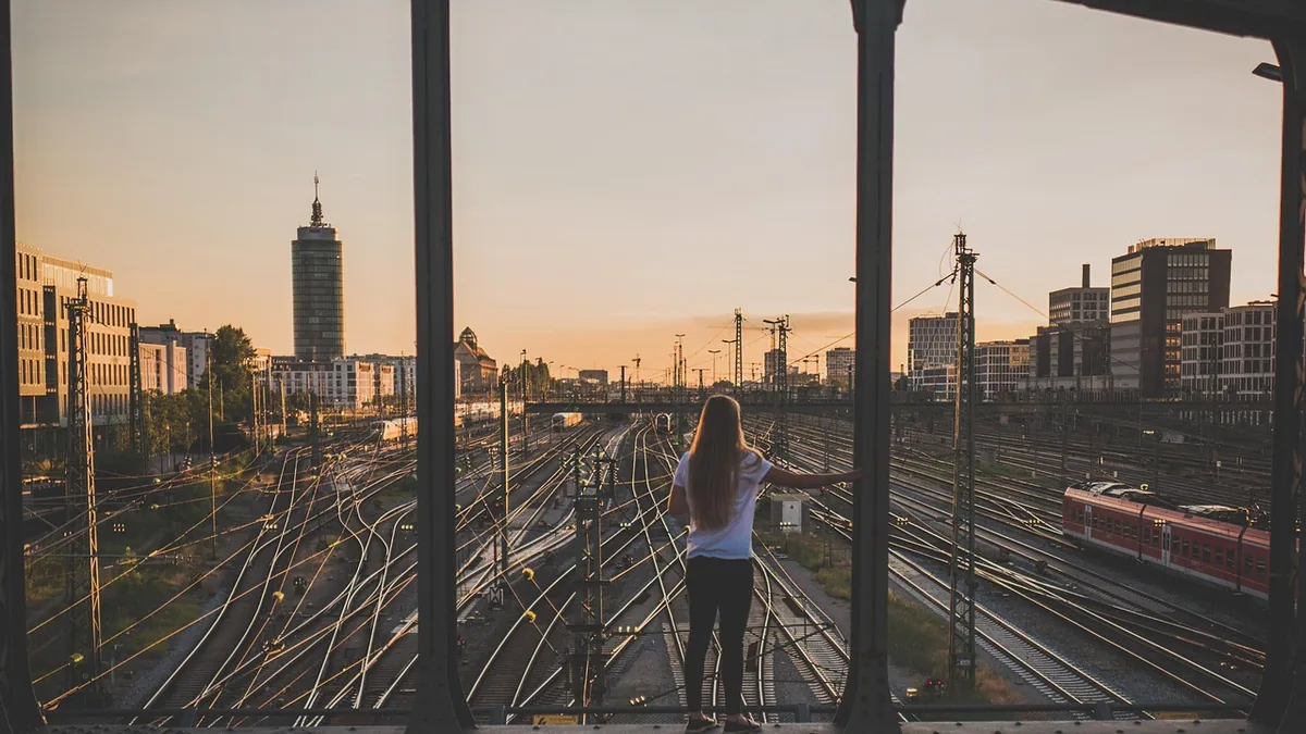 A woman overlooking a vast, complex railway network merging into a city skyline at sunset.