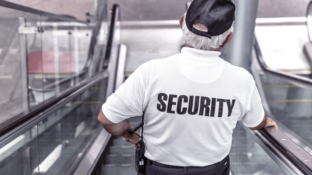 Security guard from behind wearing a white shirt with 'SECURITY' printed on it, on an escalator.