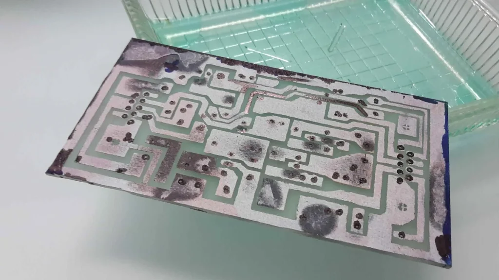 Bare printed circuit board with green and silver traces, lying next to a green glass dish.