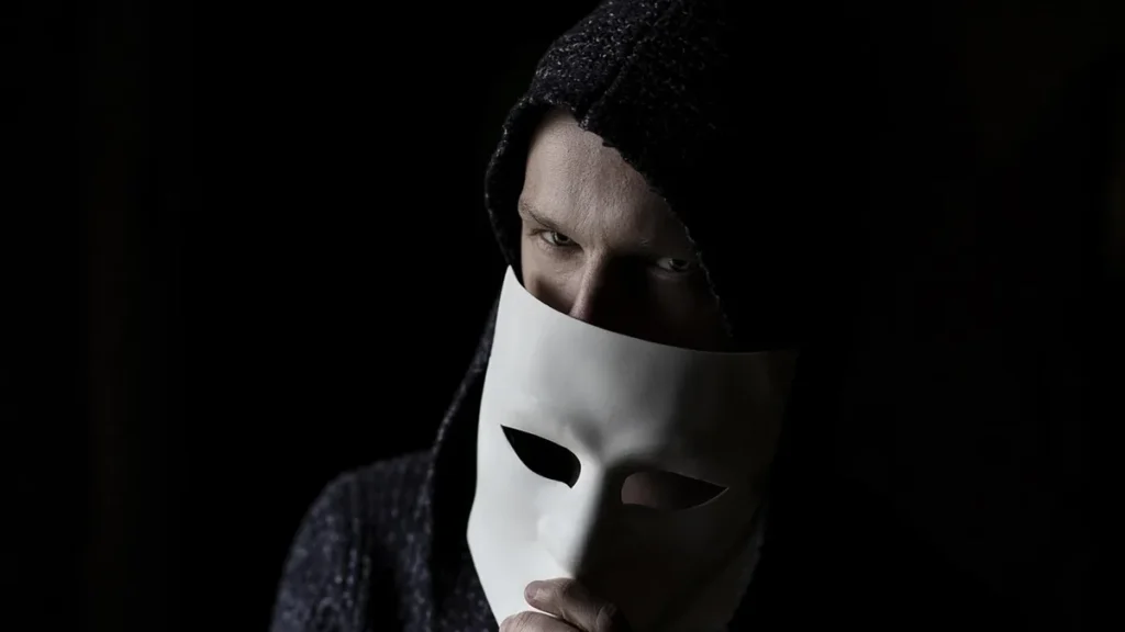 A hooded figure holding a white mask, peering intensely from the dark shadows.