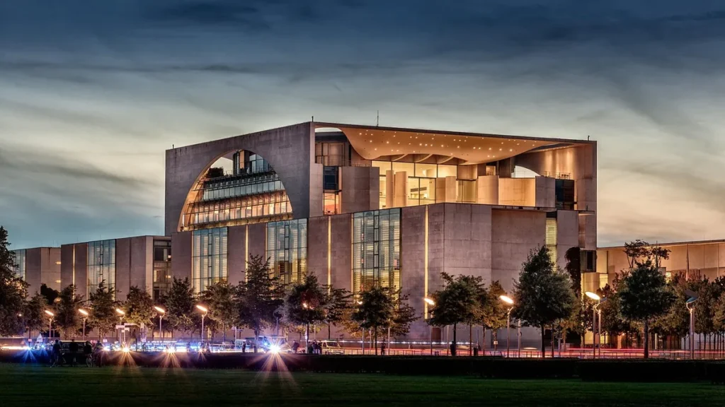 Illuminated concrete Federal Chancellery building in Berlin at dusk, with trees and streetlights.