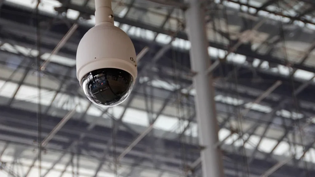 A white dome security camera hangs from a ceiling against a background of metal scaffolding.
