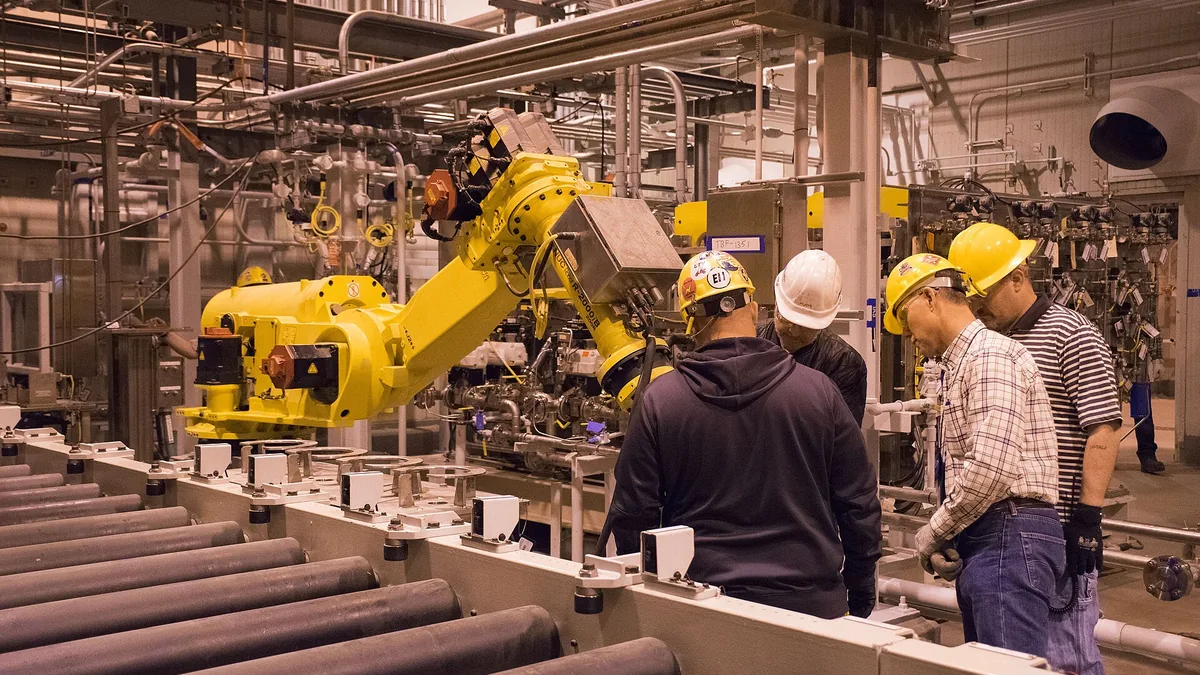 Workers in hard hats inspect a yellow industrial robotic arm on a factory production line.