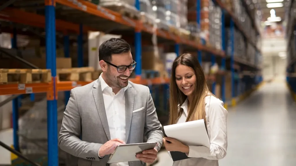 Two business professionals smiling while reviewing data on a tablet in a warehouse corridor.