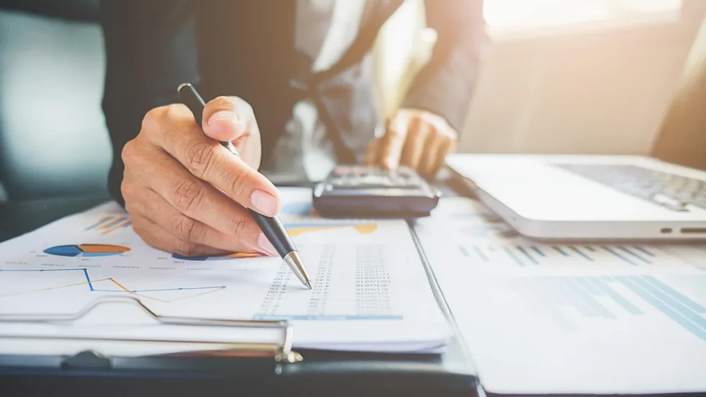 Hand holding a pen pointing at a data table on a clipboard, with charts, calculator, and laptop on a desk.