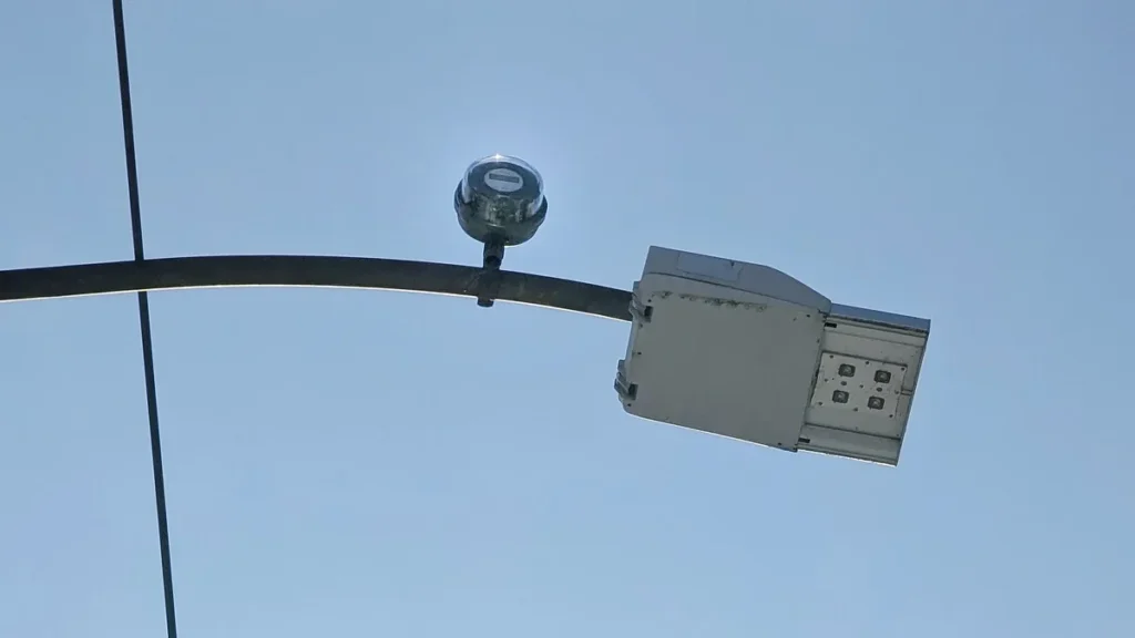 Close-up of a dome sensor mounted on a metal pole arm next to a modern LED light fixture against a blue sky.