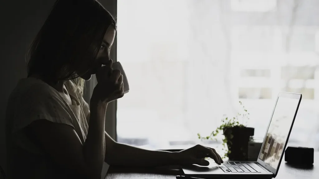 Silhouette of a woman drinking from a mug while typing on a laptop near a bright window.