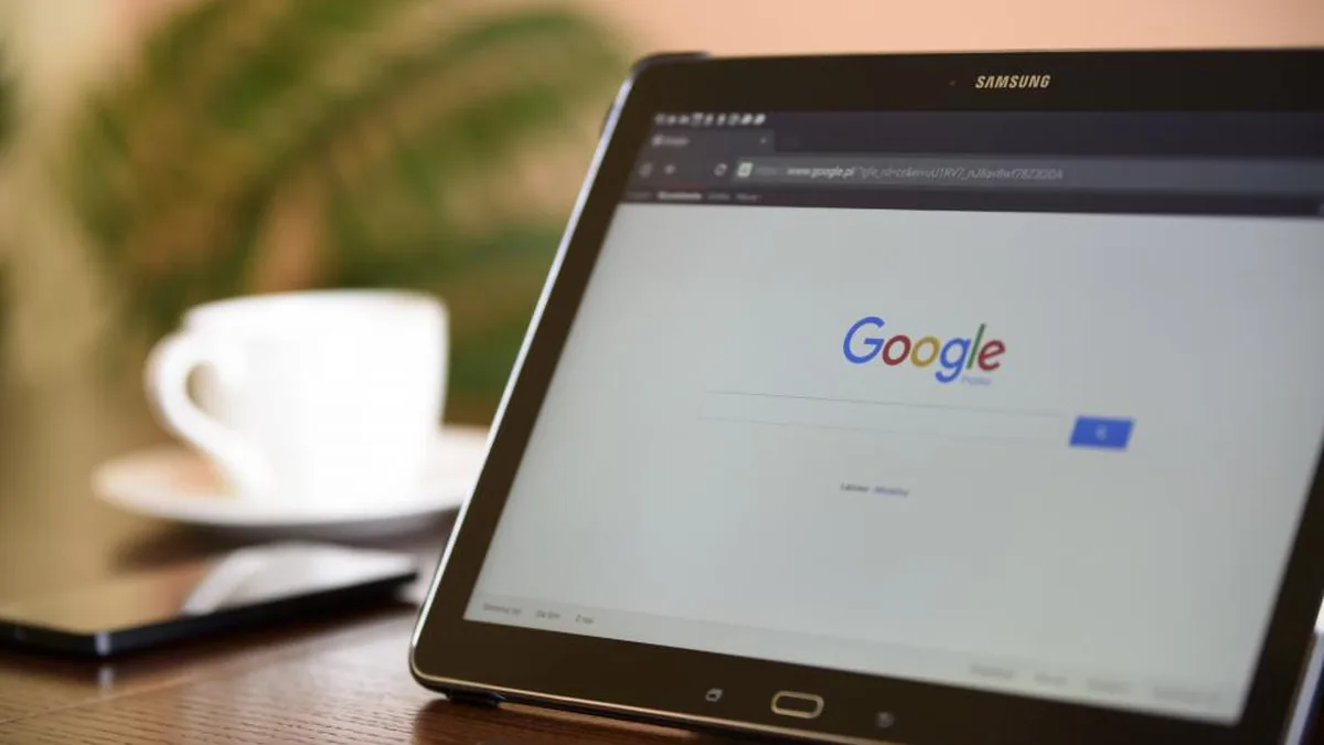 A Samsung tablet displaying the Google homepage sits on a wooden table next to a white coffee cup.