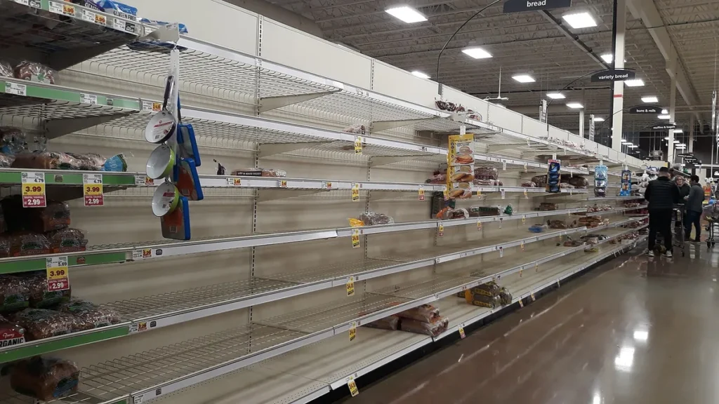 Empty shelves line a long grocery store aisle with a few shoppers in the distance.