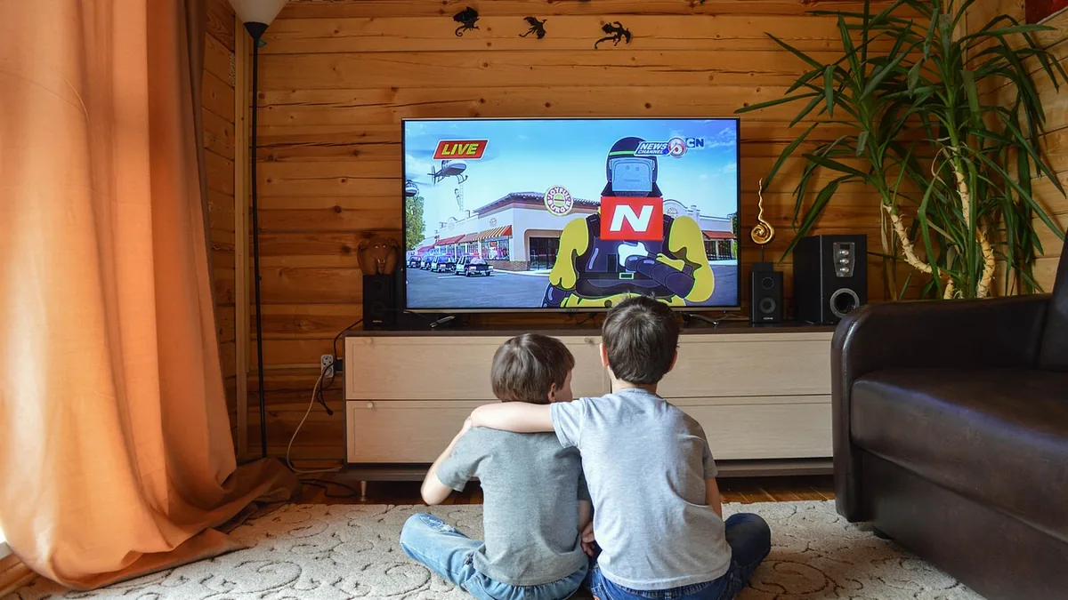 Two children sitting on the floor from behind, watching a cartoon news broadcast on a large TV screen.