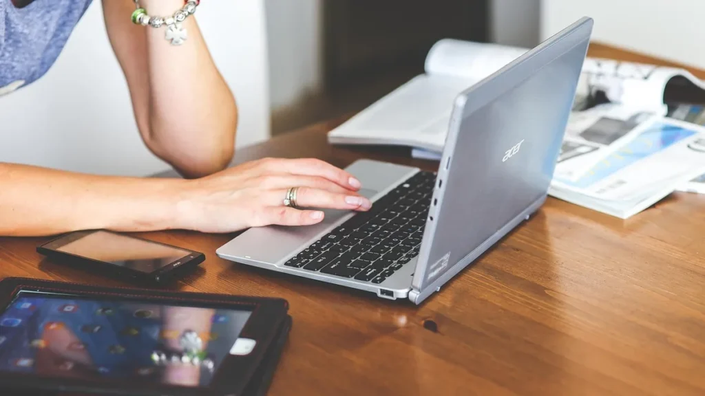 A person typing on a silver laptop on a wooden table with a smartphone and tablet nearby.