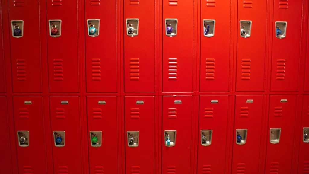 Rows of bright red metal school lockers, each secured with a colorful combination padlock in the latch.