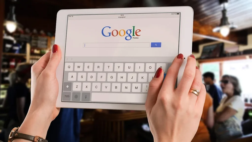 A person holding a white tablet showing the Google search homepage with an open virtual keyboard.