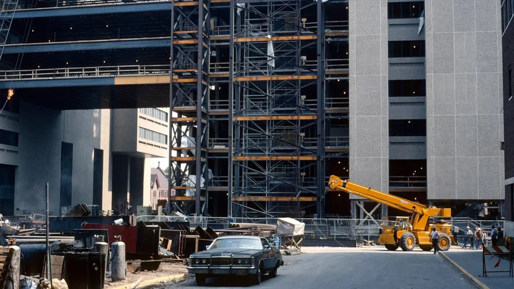 A large concrete building under construction with a yellow crane and a vintage car parked in front.
