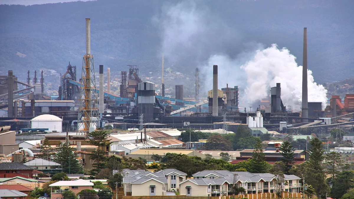 Smoking industrial chimneys loom over a residential neighborhood with hills in the background.