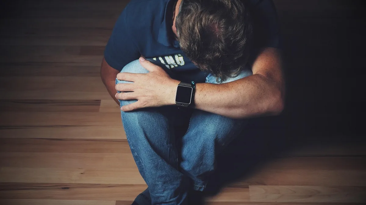 Distressed man sitting on a wooden floor with head down, holding knees, wearing a smartwatch.