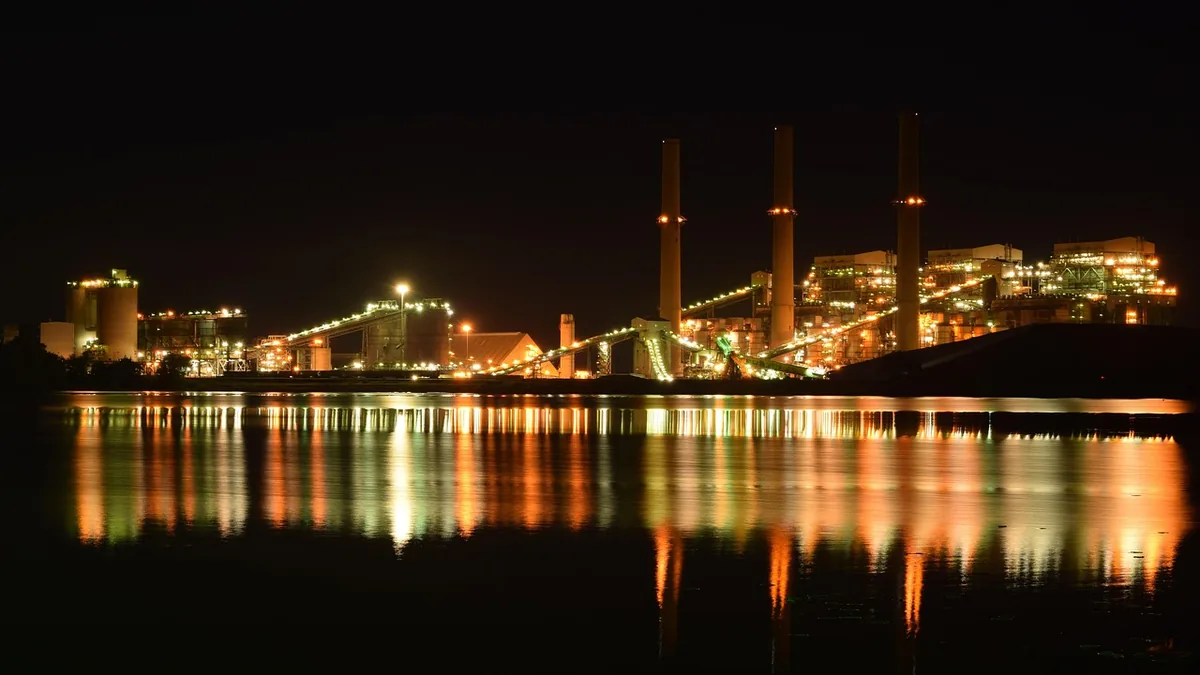 Brightly lit industrial power plant at night reflecting colorful lights across a calm dark water surface.