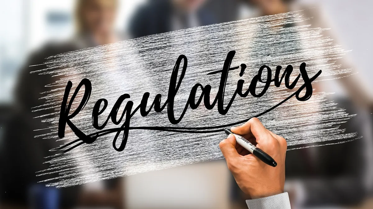 A hand writing the word 'Regulations' on a glass screen with blurred office workers in the background.
