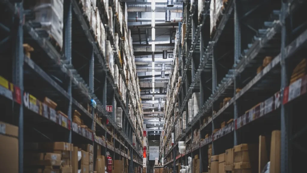 A perspective shot down a narrow aisle of tall warehouse shelves stacked with cardboard boxes.