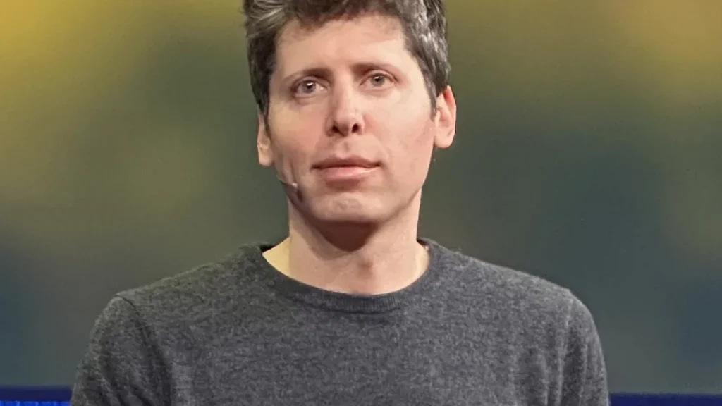 Sam Altman, CEO of OpenAI, looking forward, wearing a grey sweater against a blurred background.