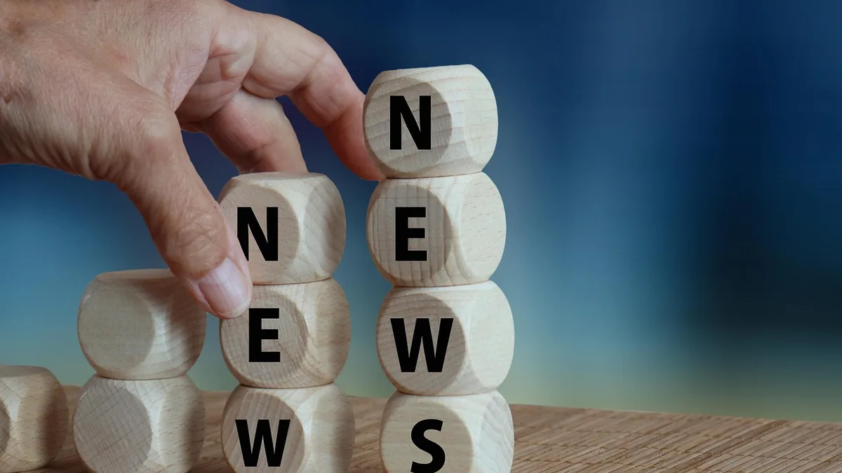 Hand arranging wooden blocks spelling NEWS on a wooden surface against a blue gradient background.