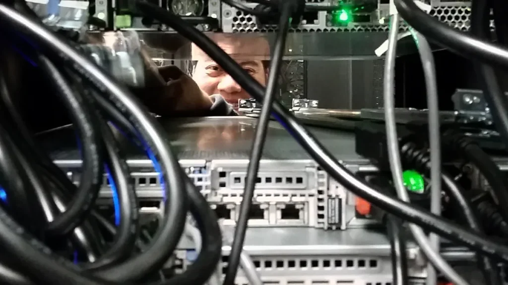 A technician's reflection peers through a dense tangle of black cables and silver server hardware inside a data center rack.